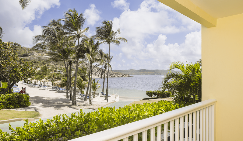 Club Room Garden View balcony overlooking tall palm trees, lush green foliage, white sand beach, and the ocean bay at St James’s Club & Villas by Kenwood Travel