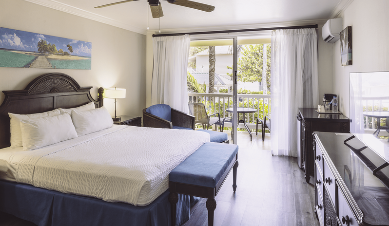Club Room Garden View king-size bed with blue accents, dark wood furniture, and sliding glass doors opening to a private patio at St James’s Club & Villas by Kenwood Travel