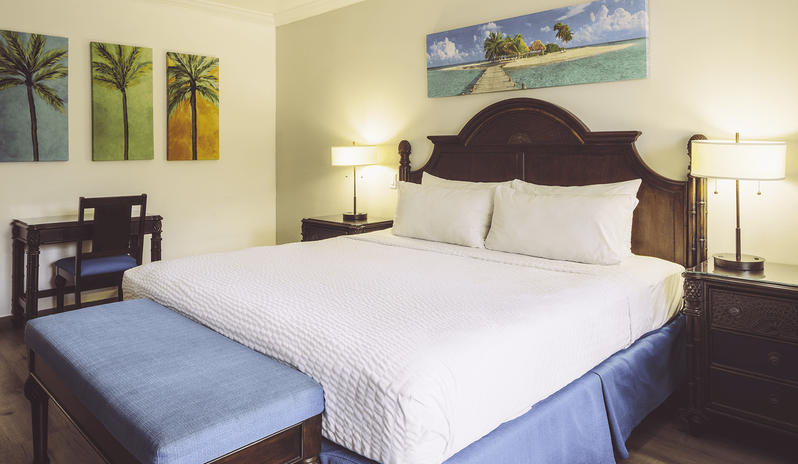 Club Room Garden View king-size bed, dark carved wood headboard, blue bed bench, bedside lamps, and a small study desk with chair at St James’s Club & Villas by Kenwood Travel