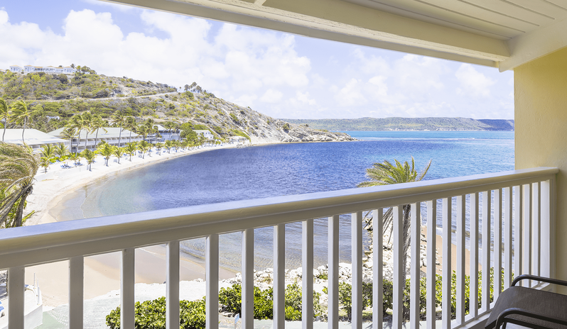 Premium Oceanview Partial Ocean View Private balcony of a premium hotel room offering an elevated panoramic view of the sandy beach bay, tropical palm trees, and green hillside at St James’s Club & Villas by Kenwood Travel