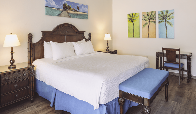 Premium Oceanview Partial Ocean View carved wood headboard, bedside tables with lamps, blue upholstered bench, and writing desk with chair at St James’s Club & Villas by Kenwood Travel