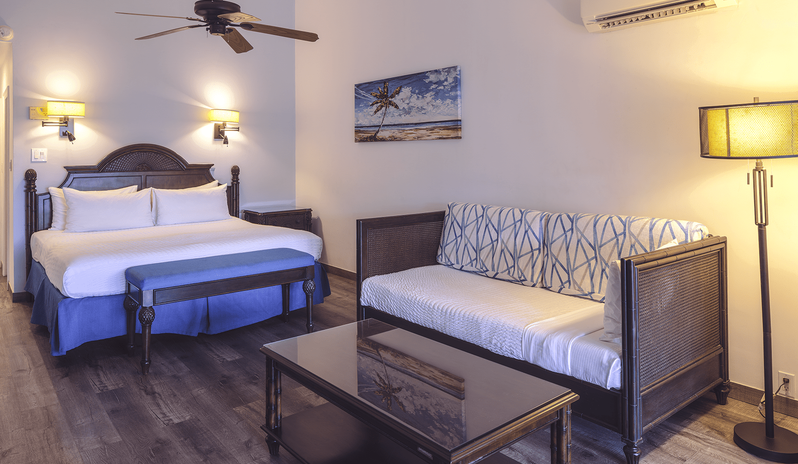 Junior Suite Ocean Facing king-size bed, blue cushioned bench, dark wood coffee table, and a patterned sofa under warm ambient lighting at St James’s Club & Villas by Kenwood Travel