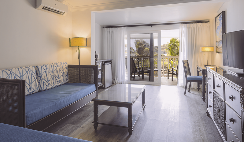 Junior Suite Ocean Facing Hotel suite living area with a blue daybed, glass coffee table, TV console, and sliding glass doors opening to a private balcony surrounded by palm trees at St James’s Club & Villas by Kenwood Travel