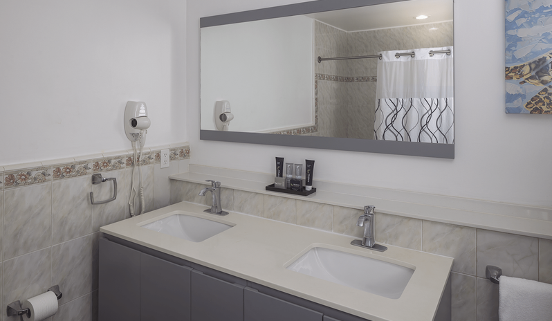Junior Suite Ocean Facing bathroom with a grey double-sink vanity, large framed mirror, wall-mounted hairdryer, and complimentary toiletries at St James’s Club & Villas by Kenwood Travel