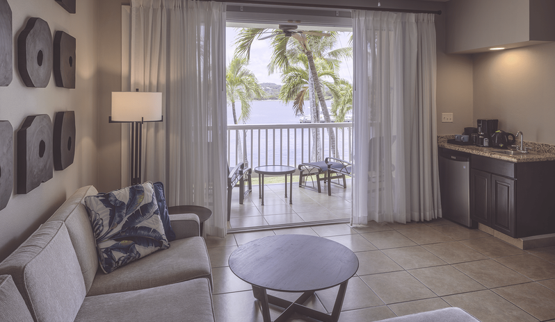 Royal Suite Bay View living area with a light grey sectional sofa, round wooden coffee table, wall art decor, and sliding glass doors opening to a covered patio with lounge chairs at St James’s Club & Villas by Kenwood Travel