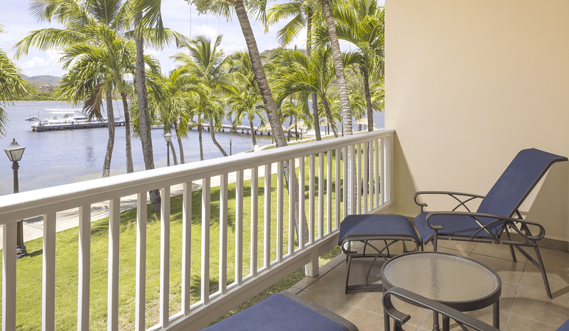 Royal Suite Bay View navy blue lounge chairs, footrests, and a round glass drinks table overlooking tropical palm trees, a green lawn, and the bay at St James’s Club & Villas by Kenwood Travel