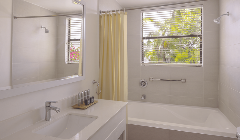 Two Bedroom Villa bathroom featuring a deep soaking bathtub with shower, white vanity sink, complimentary toiletries, and a window overlooking tropical foliage at St James’s Club & Villas by Kenwood Travel