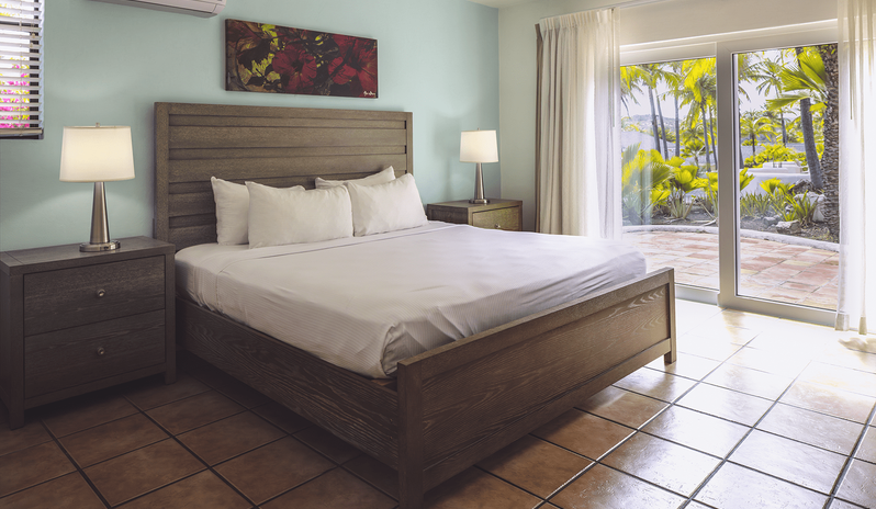 Two Bedroom Villa wooden king bed, light blue accent wall, tiled flooring, and sliding glass doors opening to a garden patio with palm trees at St James’s Club & Villas by Kenwood Travel