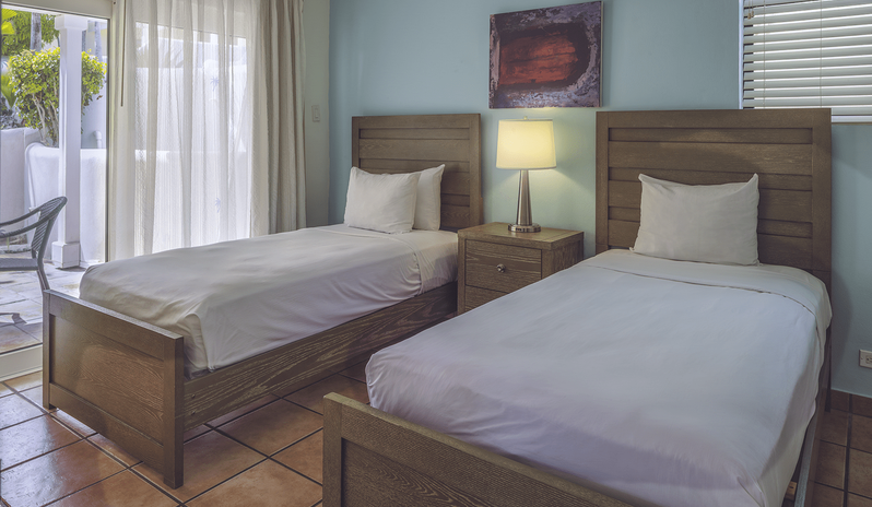 Two Bedroom Villa bedroom featuring two twin wooden beds with white linens, a bedside table lamp, and sliding glass doors leading to an outdoor patio at St James’s Club & Villas by Kenwood Travel