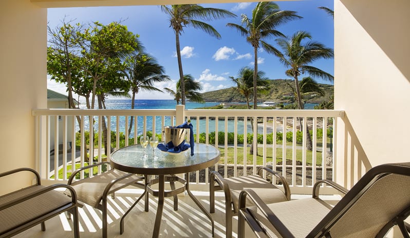 Junior Suite Ocean Facing private balcony with outdoor seating, a round glass table with champagne, and views of tropical palm trees and the ocean at St James’s Club & Villas by Kenwood Travel