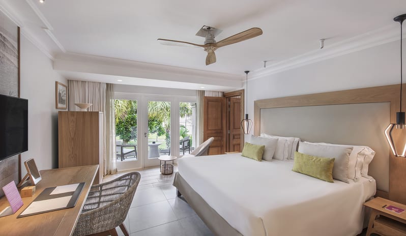 Family Room king-size bed with light green accent pillows, wooden desk with woven chair, ceiling fan, and French doors opening to an outdoor garden patio at Sugar Beach by Kenwood Travel