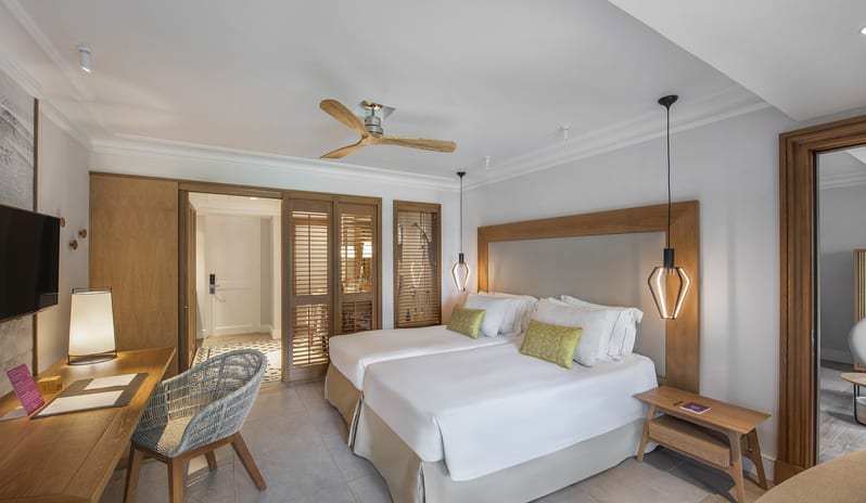Family Room two twin beds with green accent pillows, modern pendant lighting, a wooden work desk with woven chair, and wooden louvered doors at Sugar Beach by Kenwood Travel