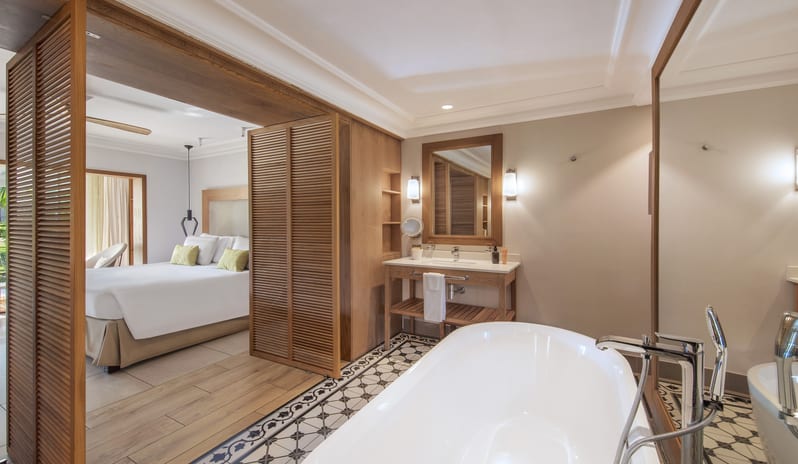 Premium Sea View Room bathroom featuring a freestanding oval soaking tub with modern chrome fixtures, patterned floor tiles, wooden vanity, and open louvered shutters leading to the bedroom at Sugar Beach by Kenwood Travel