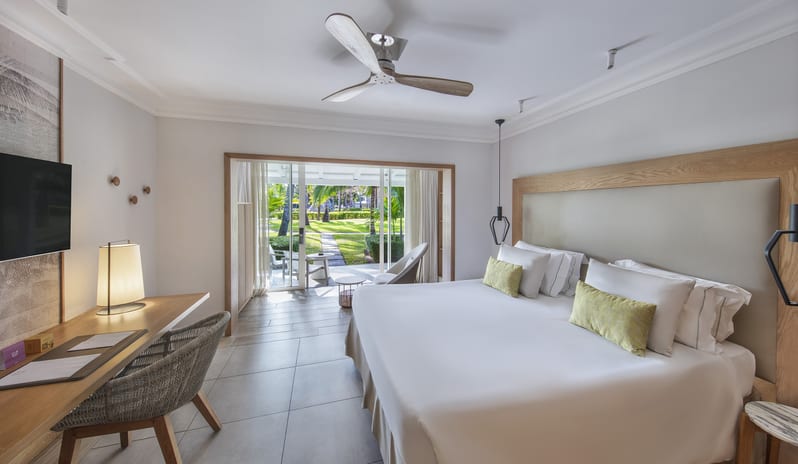 Premium Sea View Room king-size bed with light green accent pillows, wooden desk with woven chair, modern ceiling fan, and sliding glass doors opening to a lush garden patio at Sugar Beach by Kenwood Travel