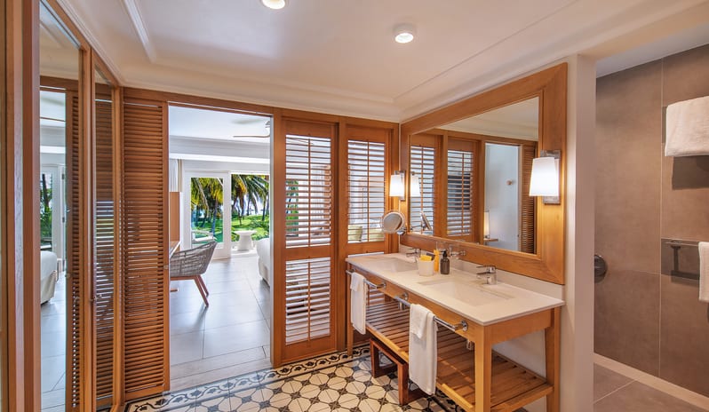 Deluxe Sea View Room bathroom featuring a wooden double vanity with dual sinks, large framed mirror, patterned tile floor, and louvered doors opening into the bedroom at Sugar Beach by Kenwood Travel