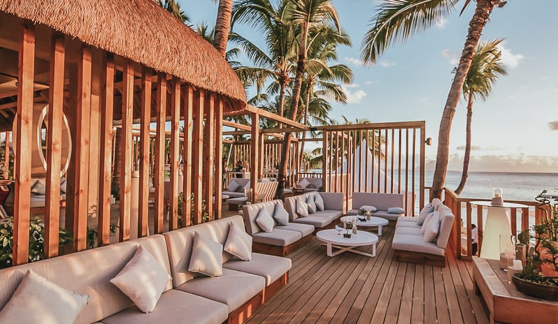 Buddha Bar Beach lounge deck with comfortable cushioned sofas, round coffee tables, tall palm trees, and sunset sky over the ocean at Sugar Beach by Kenwood Travel