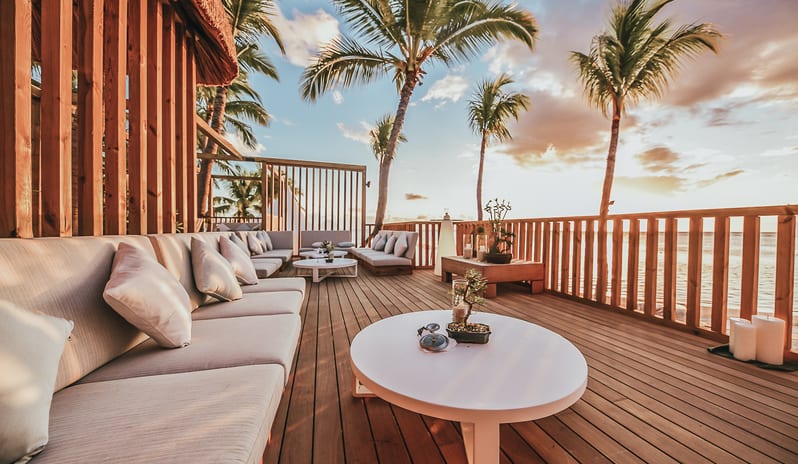 Buddha Bar Beach lounge deck with comfortable cushioned sofas, round coffee tables, tall palm trees, and sunset sky over the ocean at Sugar Beach by Kenwood Travel