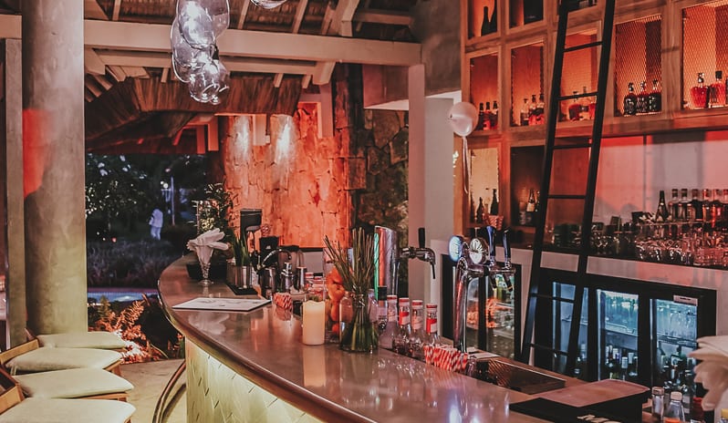 Buddha Bar Beach cocktail bar counter with bar stools, illuminated green textured front, glass pendant lights, and a warm wooden back bar display at Sugar Beach by Kenwood Travel