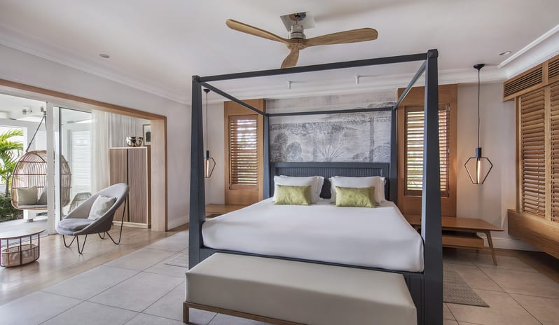 Executive Beach Front Suite modern black four-poster canopy bed, cushioned bench, geometric pendant lighting, ceiling fan, and walkout patio access with a hanging wicker egg chair at Sugar Beach by Kenwood Travel