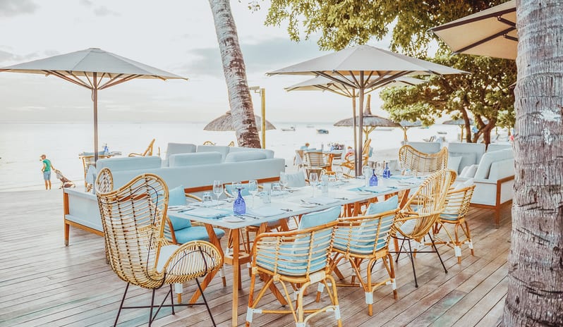 Maré Maré dining setup on a wooden deck featuring long dining tables, woven rattan chairs with light blue cushions, sun umbrellas, and ocean views at Sugar Beach by Kenwood Travel
