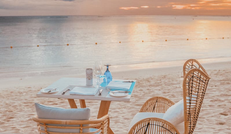 Maré Maré seaside dining table for two on a wooden deck by the sandy beach, featuring wicker chairs and tableware at sunset at Sugar Beach by Kenwood Travel