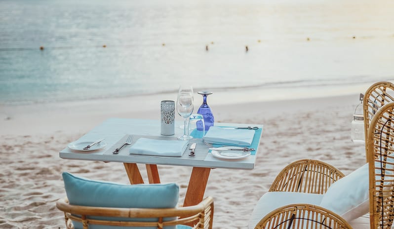 Maré Maré beachside dining table set with wine glasses, blue water goblet, folded napkins, and wicker chairs overlooking the sandy shore and ocean at Sugar Beach by Kenwood Travel