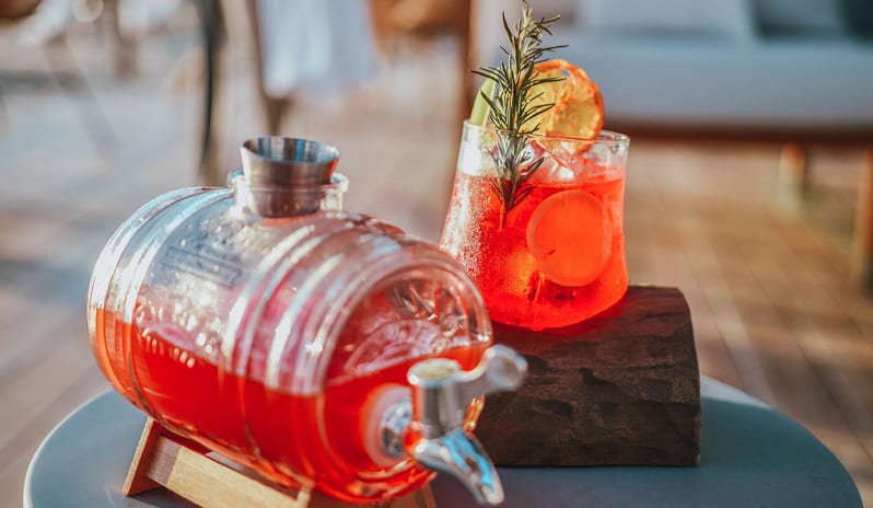 Maré Maré cocktail garnished with a fresh rosemary sprig and dehydrated orange slice, served alongside a glass mini beverage barrel dispenser on a wooden stand at Sugar Beach by Kenwood Travel